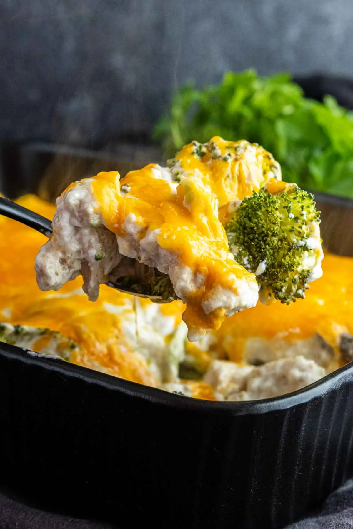 A spoon lifts a serving of high protein chicken and broccoli casserole from a black baking dish, with melted cheese visible on top—a delicious high protein meal idea.