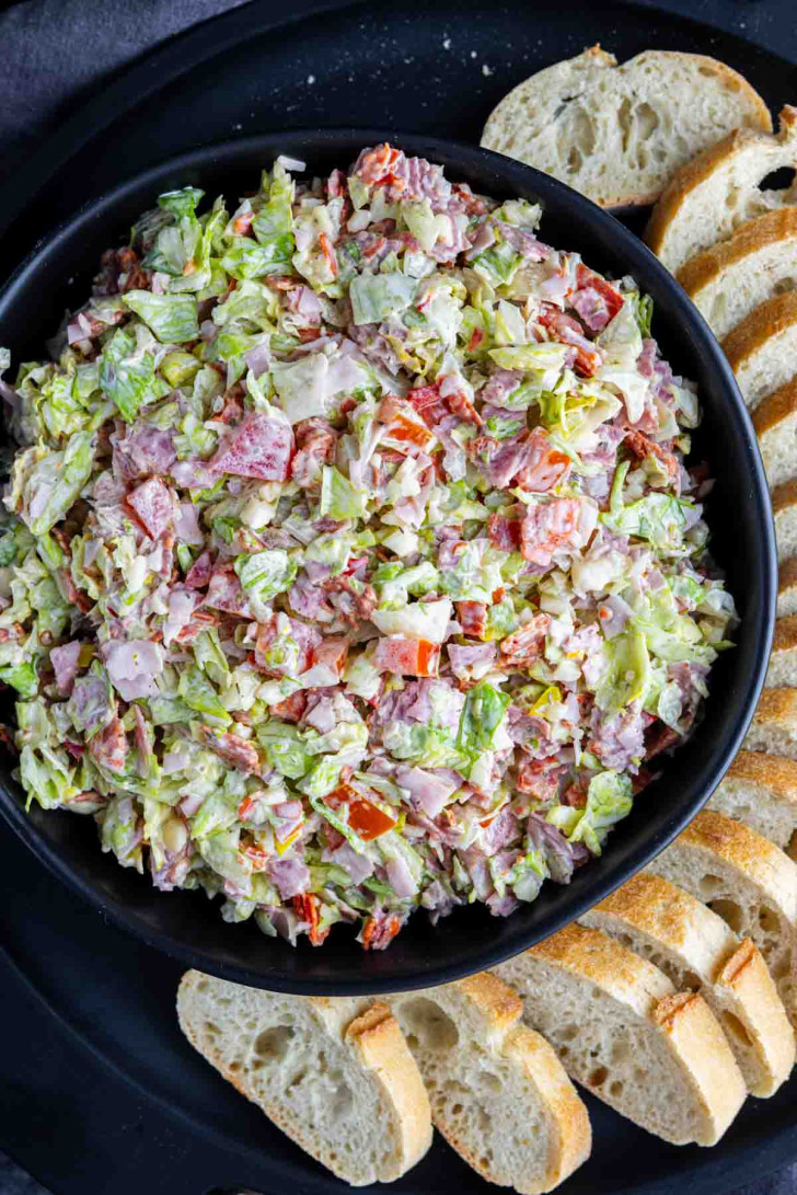 A black bowl filled with Italian chopped salad dip made of lettuce, meat, and tomatoes, surrounded by slices of baguette on a black plate.