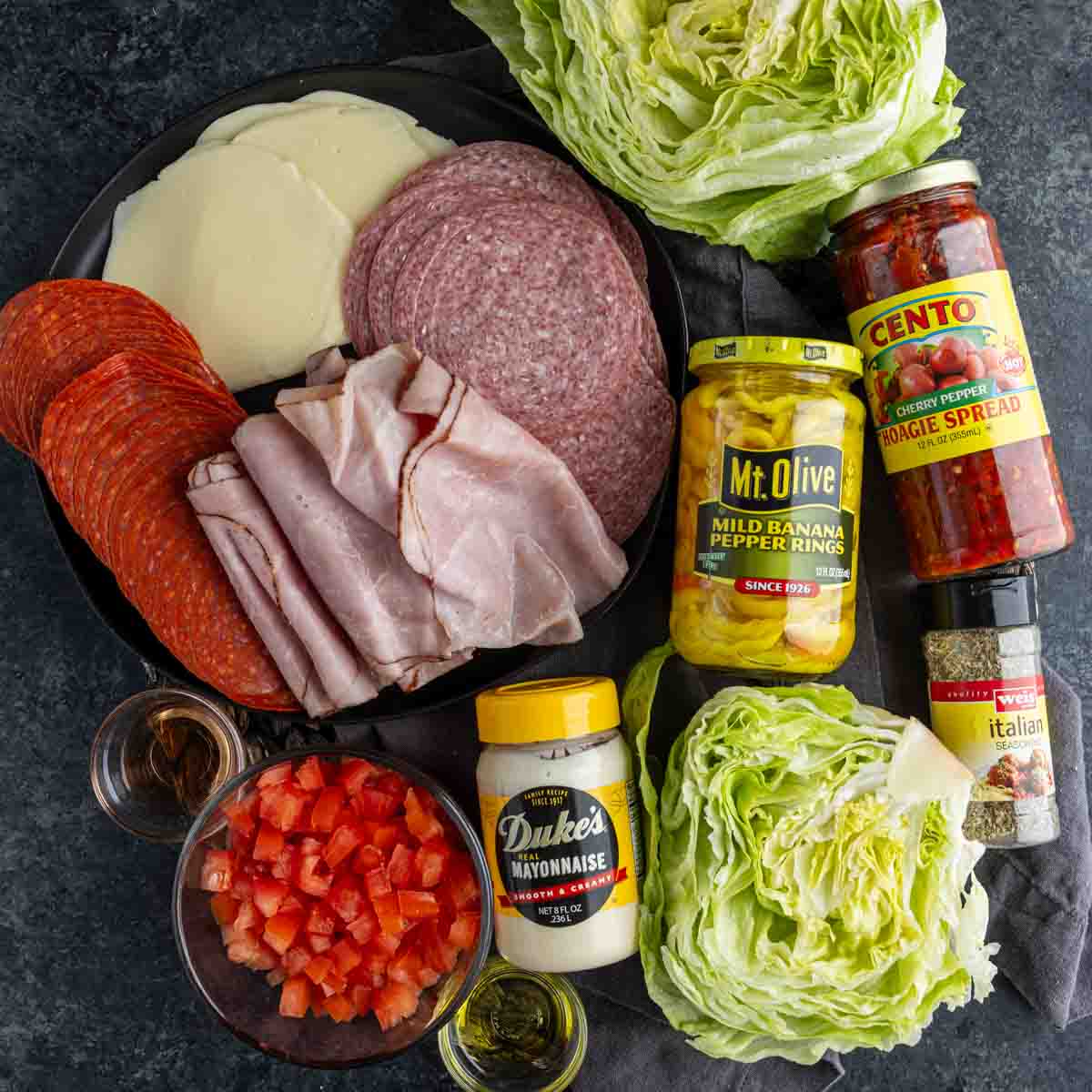 Assorted deli meats, provolone cheese, iceberg lettuce, chopped tomatoes, mayonnaise, banana pepper rings, giardiniera, Italian seasoning, and olive oil arranged for an easy hoagie dip—perfect for your next game day gathering.