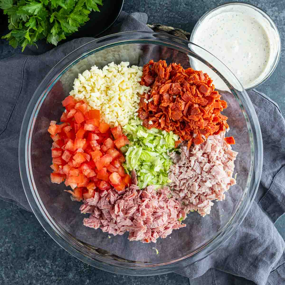A glass bowl with chopped tomatoes, lettuce, ham, turkey, mozzarella cheese, and bacon in sections—this easy hoagie dip is perfect for game day. A jar of creamy dressing sits nearby for dipping.