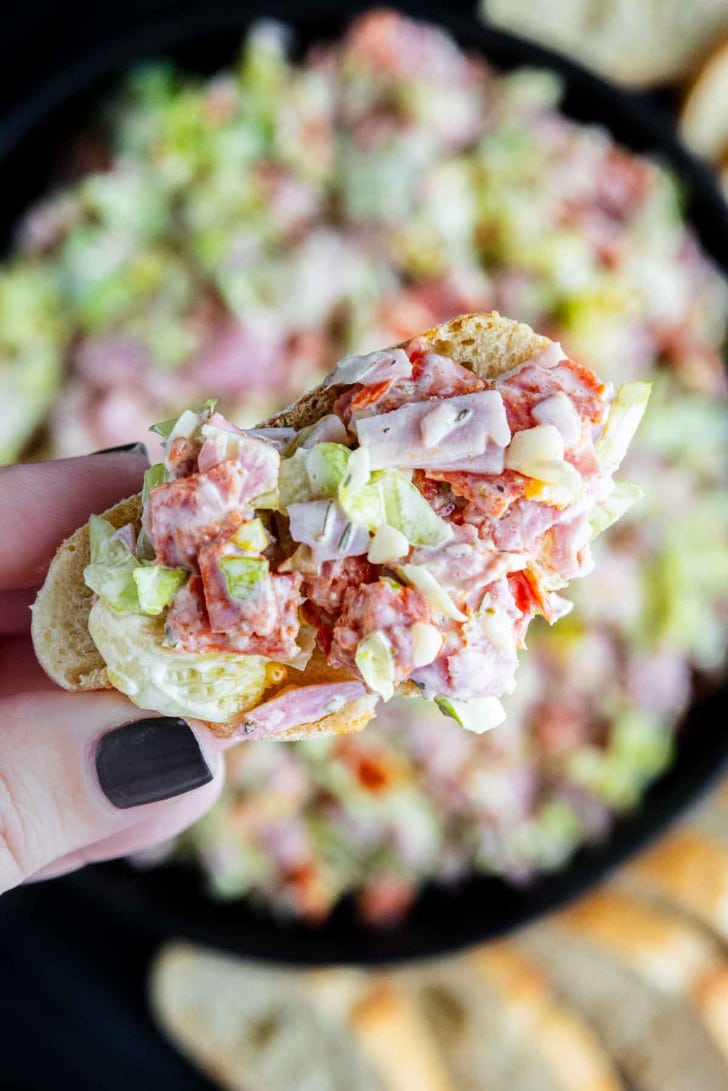 A hand holding a piece of bread topped with a chopped salad mixture of lettuce, tomato, ham, cheese, and dressing—an easy hoagie dip perfect for parties or game day snacking.