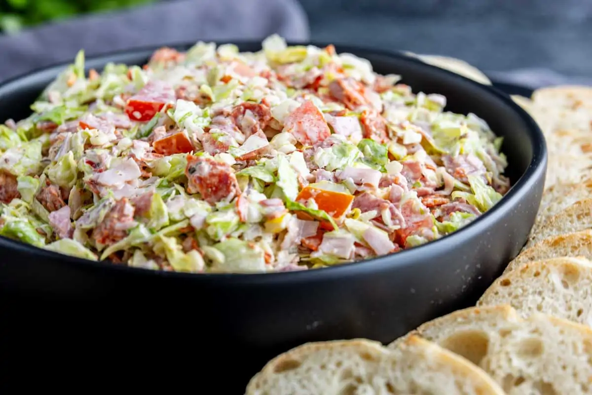 A black bowl filled with Italian chopped salad dip—lettuce, tomatoes, and deli meats—surrounded by slices of bread for dipping.