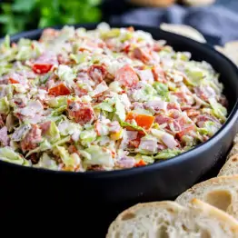 A black bowl filled with chopped salad containing lettuce, tomatoes, deli meat, and a creamy dressing, with slices of bread on the side.