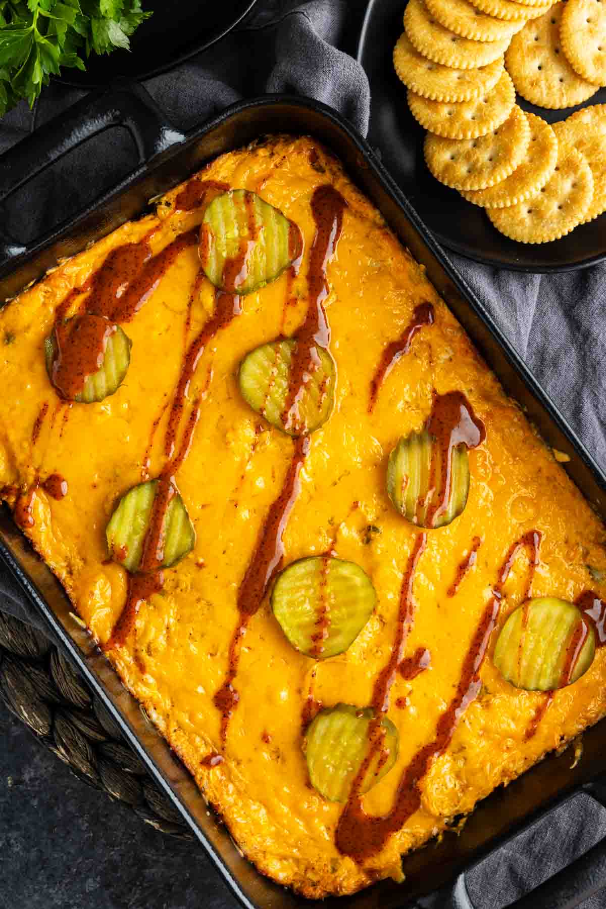 Rectangular baking dish featuring Nashville hot chicken dip with melted cheese, topped with pickle slices and a drizzle of sauce. A plate of round crackers is visible in the upper right corner.