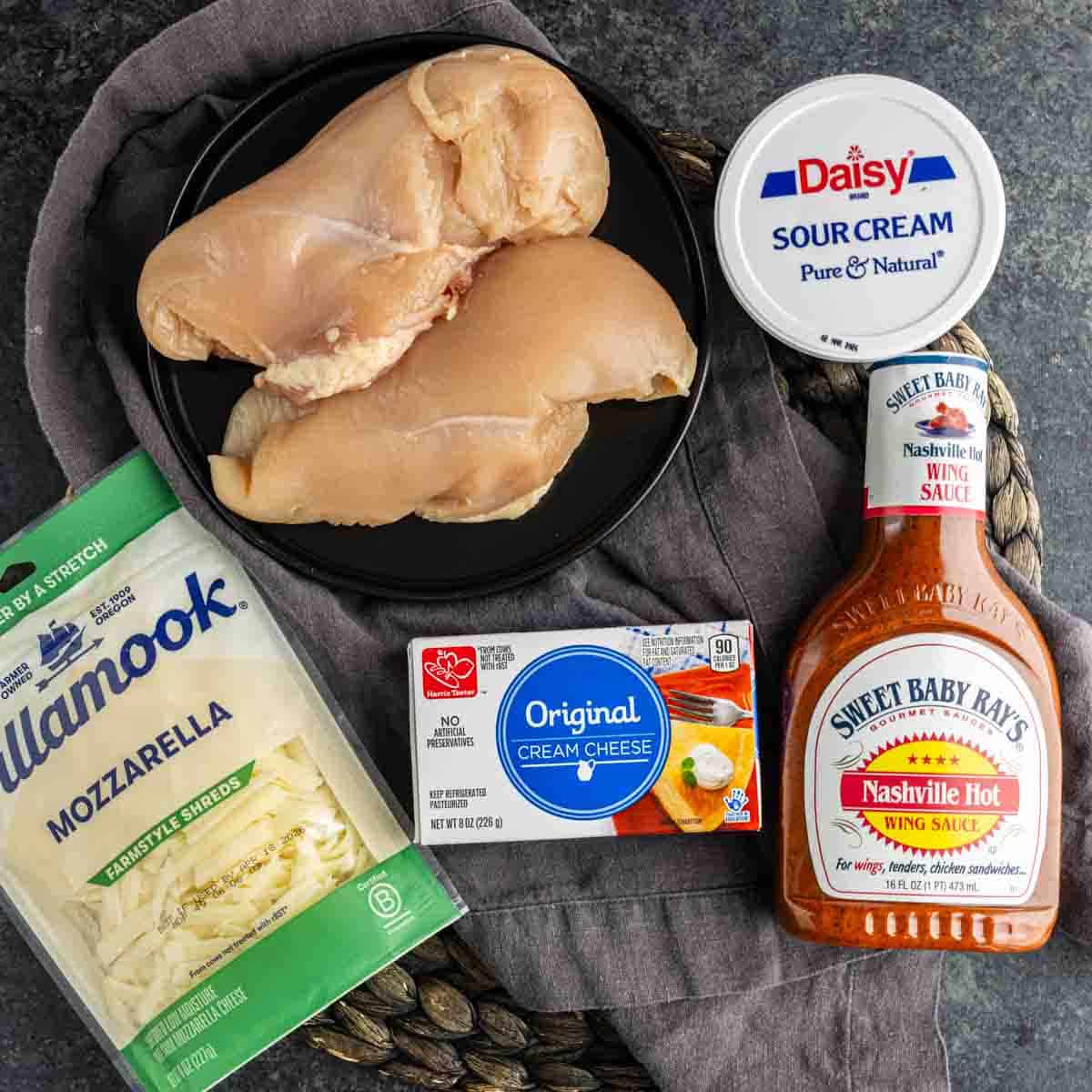 Raw chicken breasts on a black plate, with packages of mozzarella cheese, cream cheese, sour cream, and a bottle of Nashville hot wing sauce—perfect ingredients for making a hot chicken dip or party dip with shredded chicken.