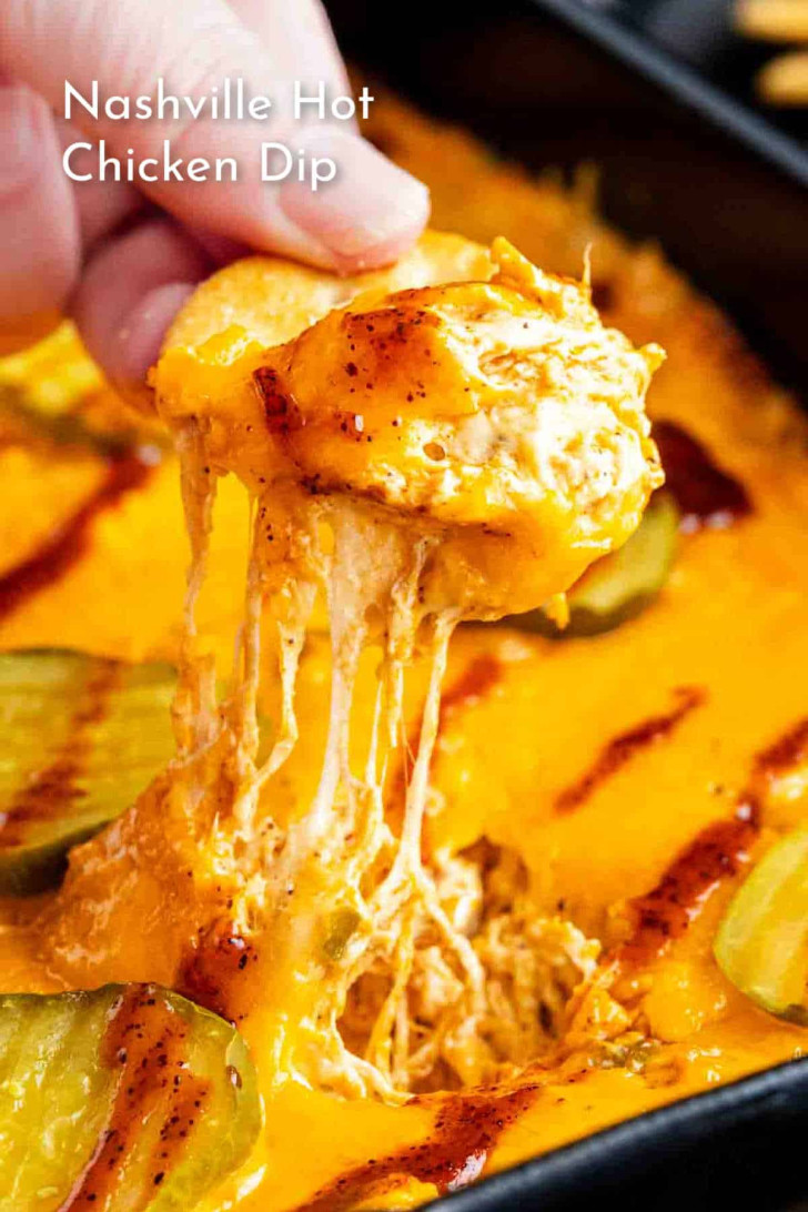 A hand dips a chip into a cheesy, baked Nashville hot chicken dip topped with melted cheese and pickles.
