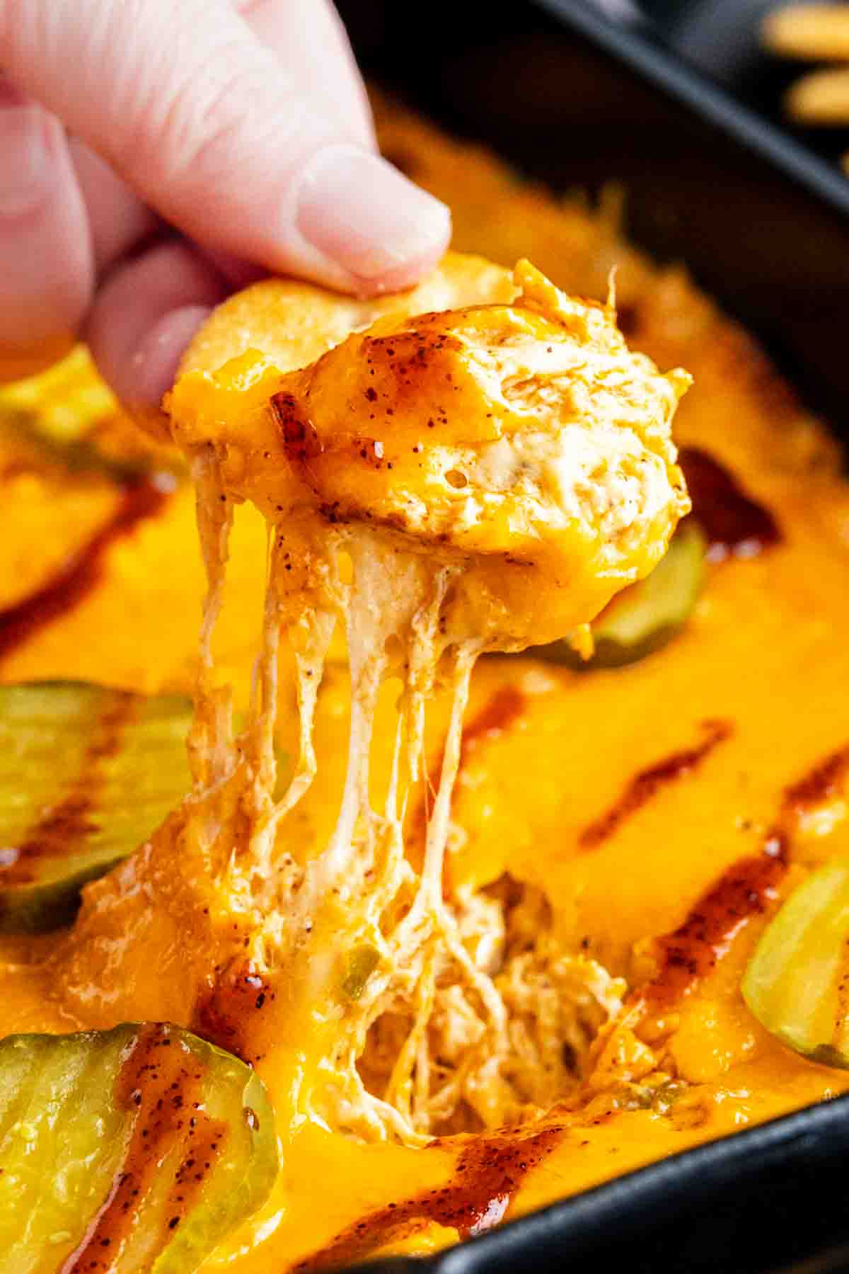A hand dipping a chip into a cheesy Nashville hot chicken dip topped with sliced pickles, showing melted cheese stretching from the dish.