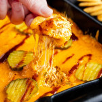 A hand pulls a cracker covered in melted, cheesy dip topped with pickles from a baking dish filled with more dip and pickle slices.