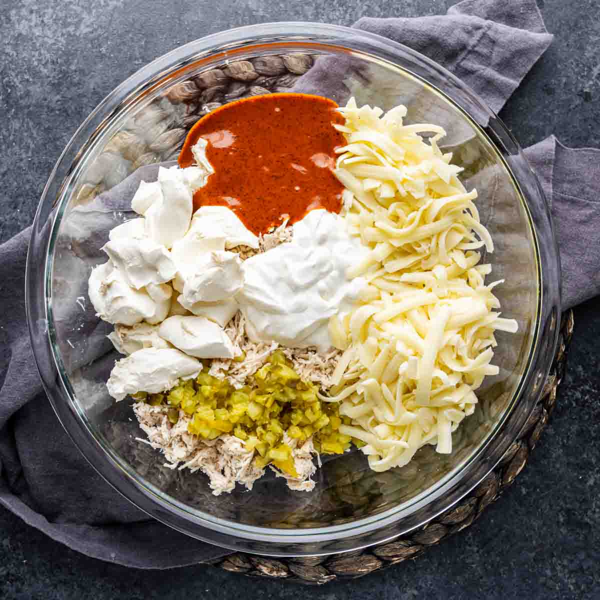 A glass bowl with shredded chicken, chopped pickles, shredded cheese, sour cream, cream cheese, and a reddish sauce—perfect as a game day dip—sits on a gray napkin atop a dark surface.