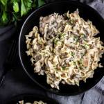 A black bowl filled with creamy beef stroganoff noodles, garnished with chopped parsley, sits on a dark cloth next to a fork and fresh parsley.
