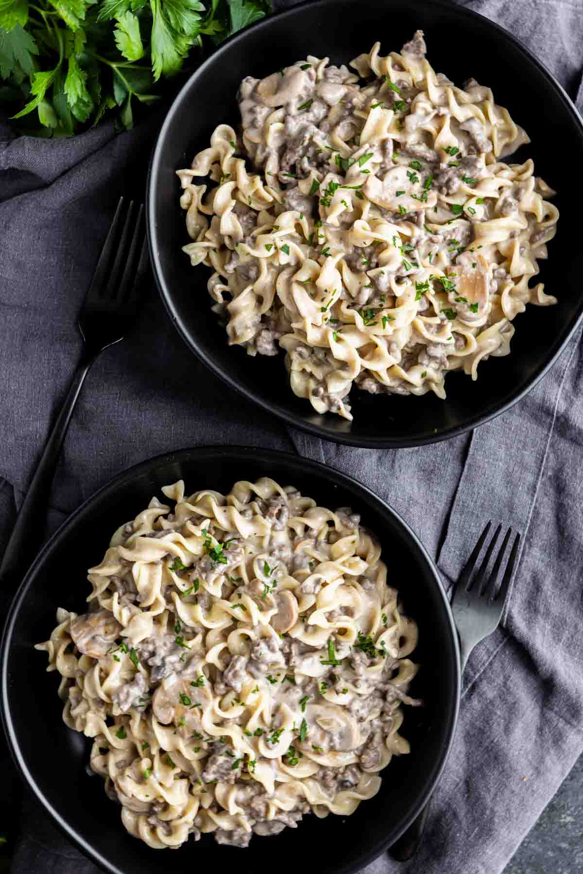 Two black bowls filled with creamy beef stroganoff pasta garnished with chopped herbs, placed on a dark cloth with two black forks beside them.