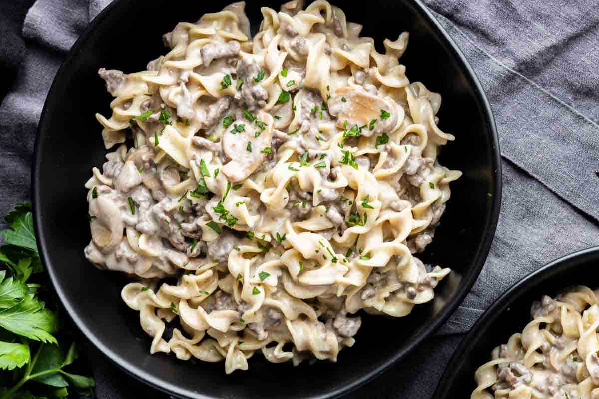 A black bowl filled with creamy beef stroganoff pasta garnished with chopped parsley, set on a dark cloth background.