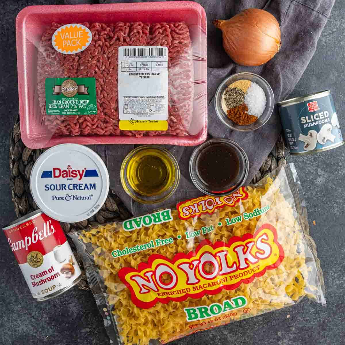 Ingredients for a beef stroganoff dish laid out, including ground beef, sour cream, egg noodles, broth, onion, seasonings, mushroom soup, mushrooms, and oil.