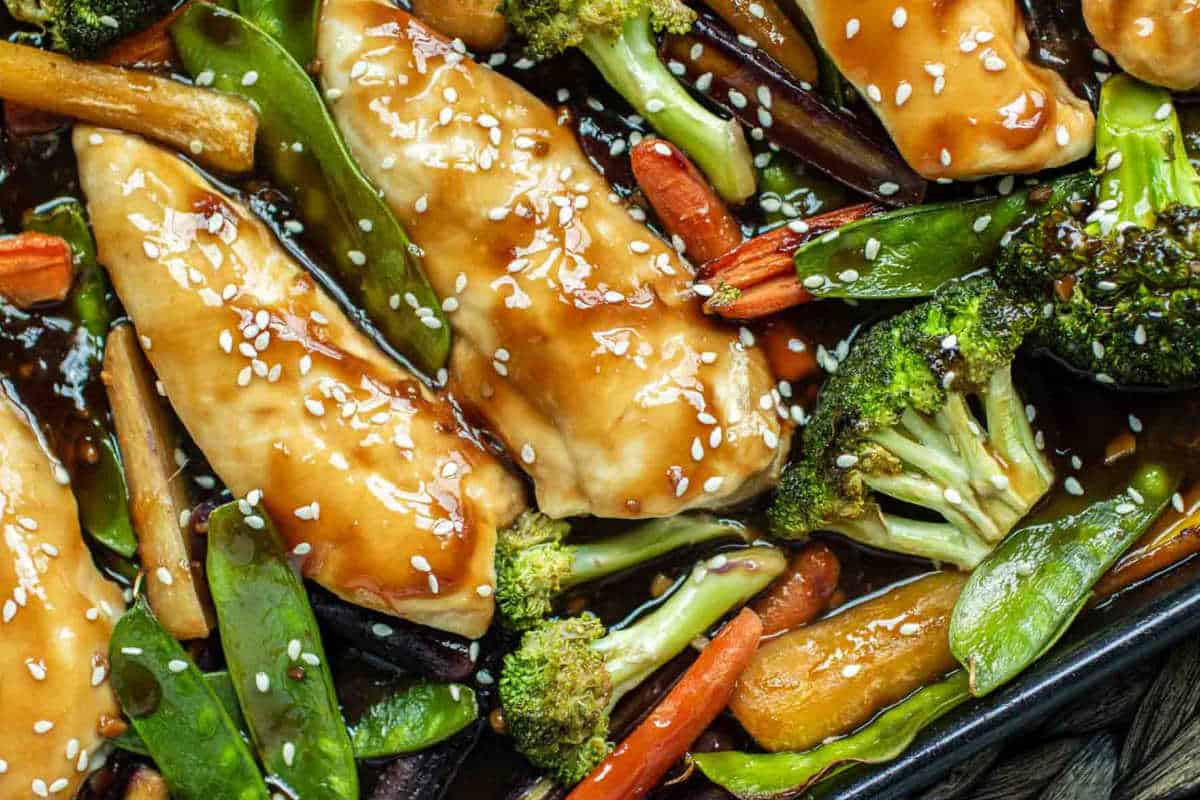 Enjoy one of the best high-protein recipes with chicken breasts glazed in teriyaki sauce, topped with sesame seeds, and served alongside fresh broccoli, carrots, and snap peas—a flavorful addition to your new year recipes collection.