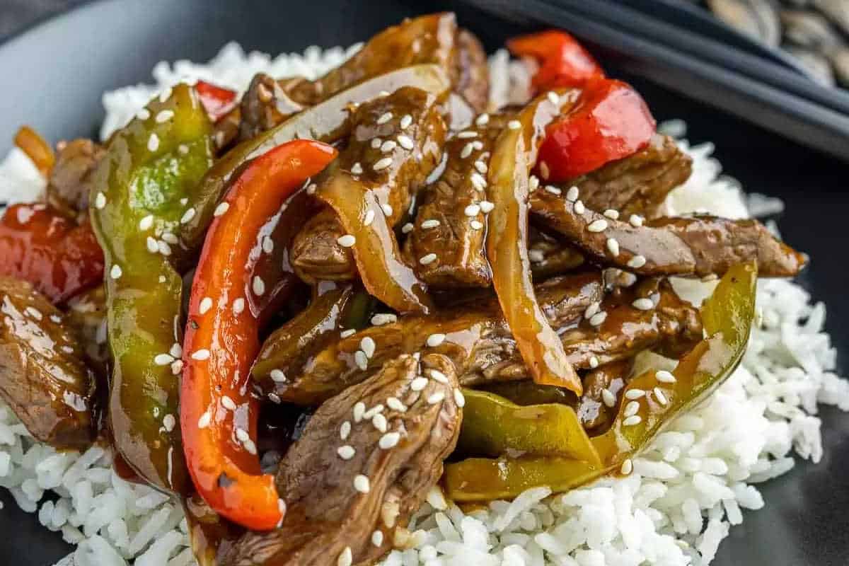 A plate of white rice topped with stir-fried beef, red and green bell peppers, onions, brown sauce, and sprinkled with sesame seeds—one of the best high-protein recipes for a satisfying and flavorful meal.