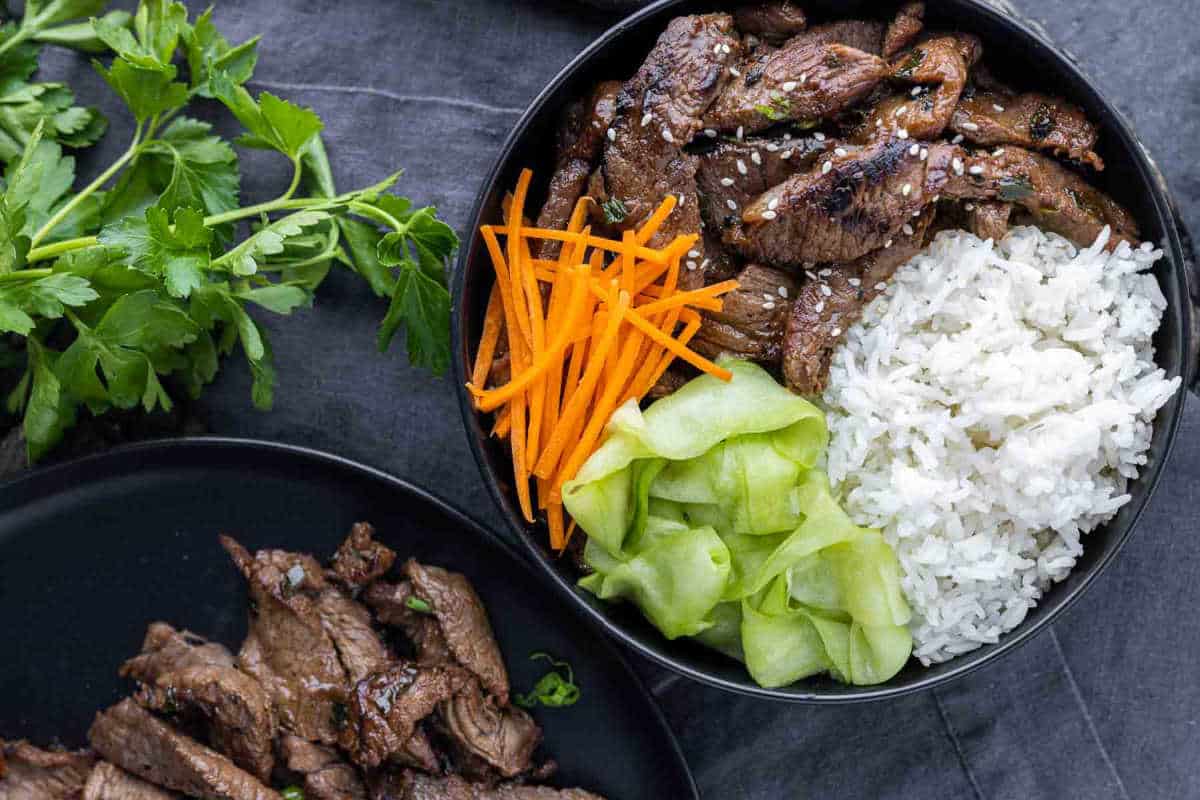 A bowl with sliced grilled beef, white rice, shredded carrots, and cucumber ribbons, garnished with sesame seeds, sits next to a plate of additional beef—one of the best recipes for high-protein meals to start the New Year right.