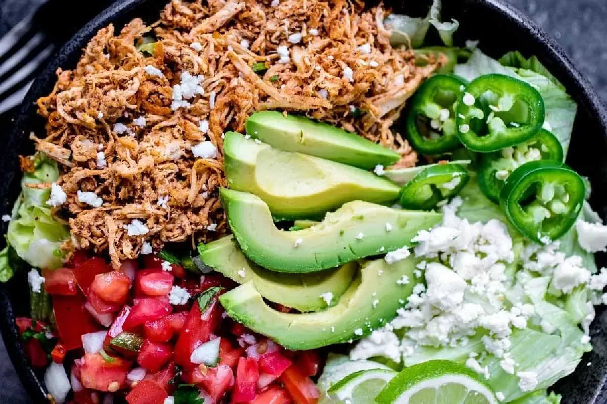 A bowl containing shredded chicken, sliced avocado, jalapeños, pico de gallo, crumbled cheese, lettuce, and lime wedges—one of the best high-protein recipes to add to your new year recipes collection.
