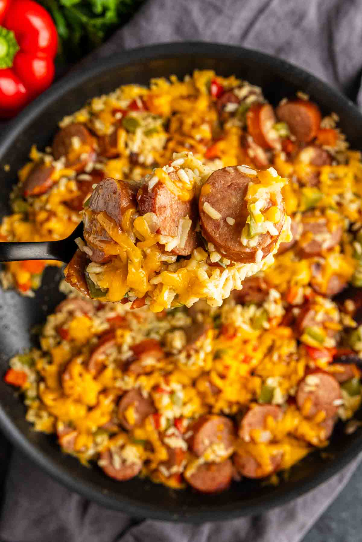 A close-up of a one pot cheesy sausage and rice skillet filled with sausage, rice, bell peppers, and melted cheese, with a spoon lifting a portion from the pan.