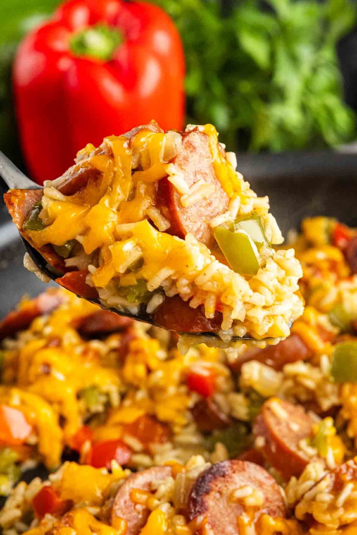 A spoonful of cheesy sausage and rice with savory sausage, bell peppers, and melted cheddar, with a red bell pepper and greens in the background.