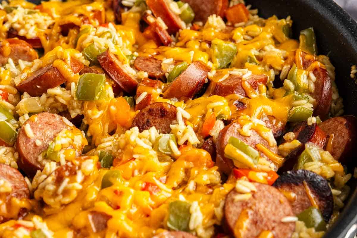 A close-up of a skillet containing sliced sausage, rice, diced green peppers, and melted cheddar cheese—an irresistible one pot cheesy sausage and rice dish.