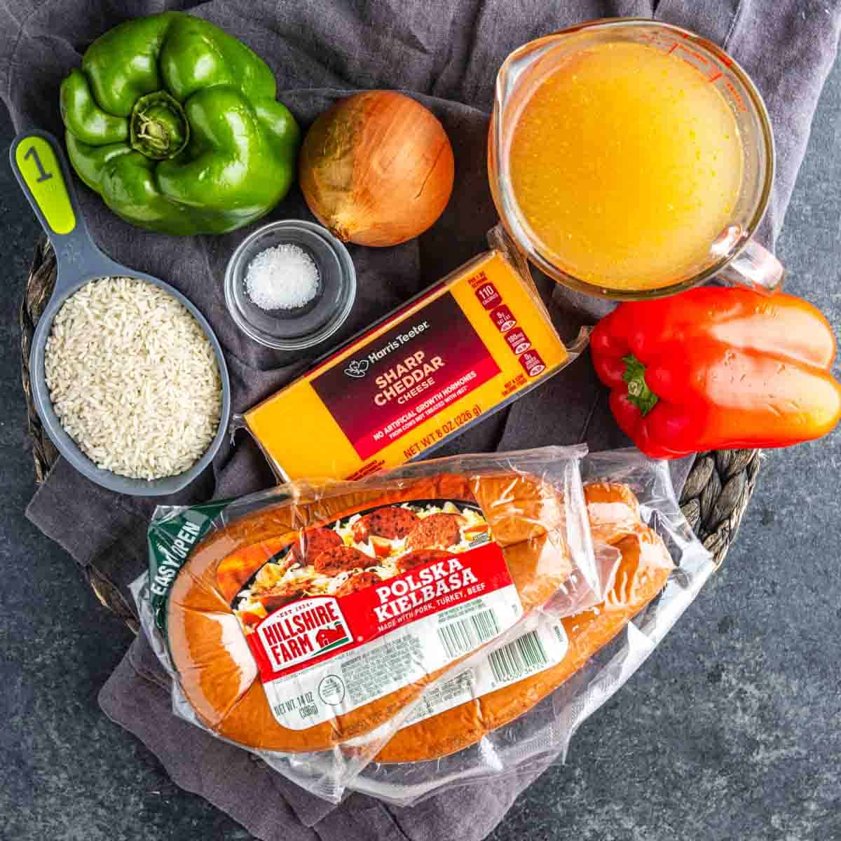 Ingredients for one pot cheesy sausage and rice on a dark surface: green bell pepper, yellow onion, salt, white rice, cheddar cheese, chicken broth, red bell pepper, and Polska kielbasa sausage.