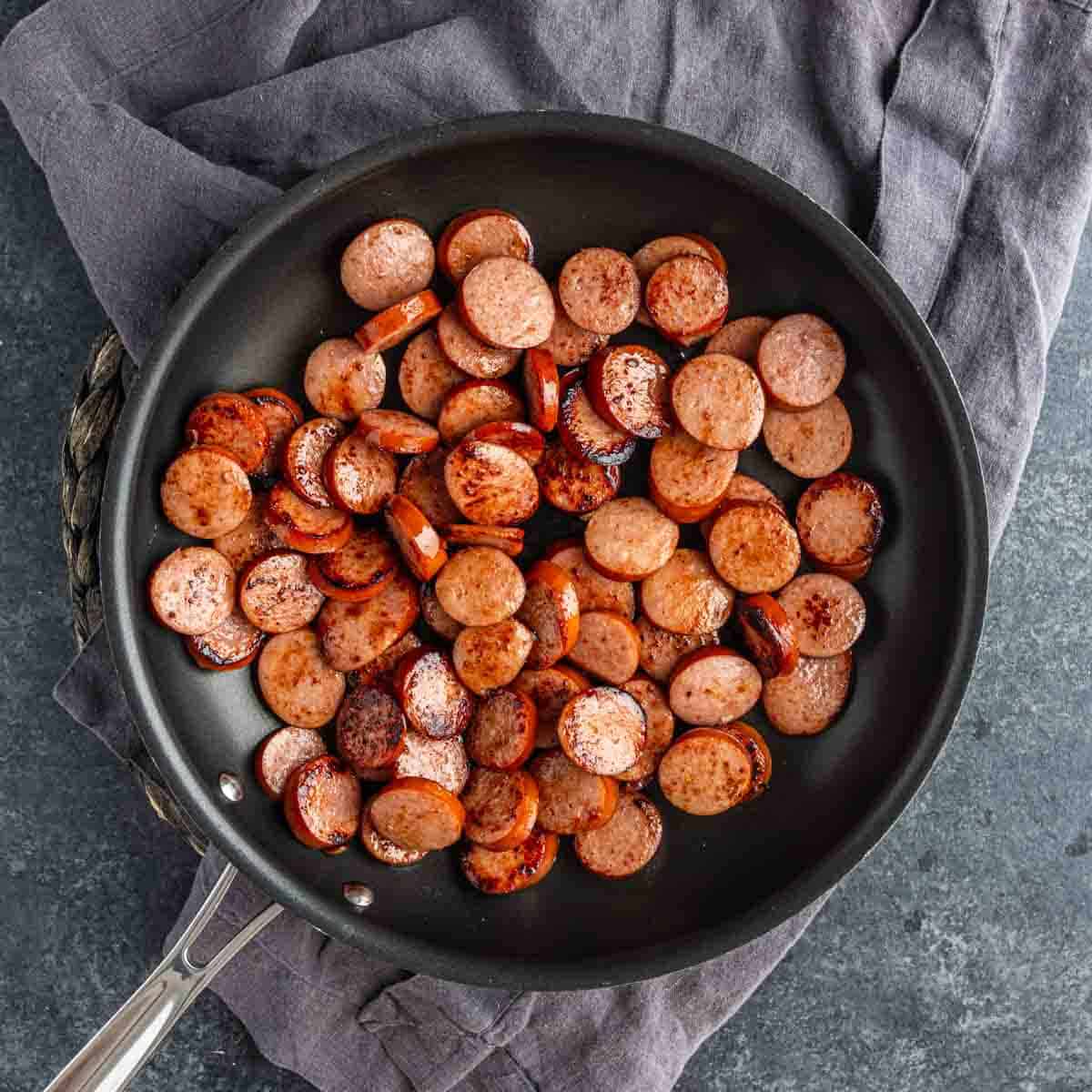 Sliced sausage pieces being browned in a black frying pan, perfect for starting a cheesy sausage and rice dish, with the pan resting on a gray cloth.