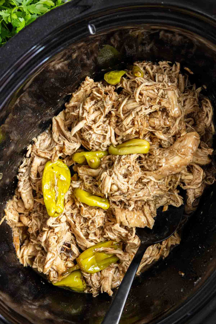 Shredded beef with pepperoncini peppers in a slow cooker, with a black serving spoon visible.
