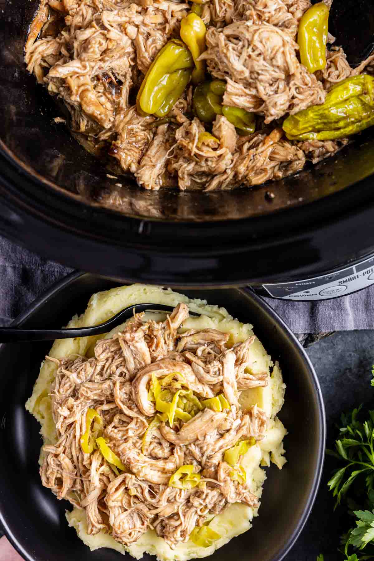 Slow Cooker Mississippi Chicken—tender, pepperoncini chicken slow-cooked in a crockpot—served over mashed potatoes in a black bowl with a fork. It's the ultimate dump and go crockpot meal for cozy comfort food nights.