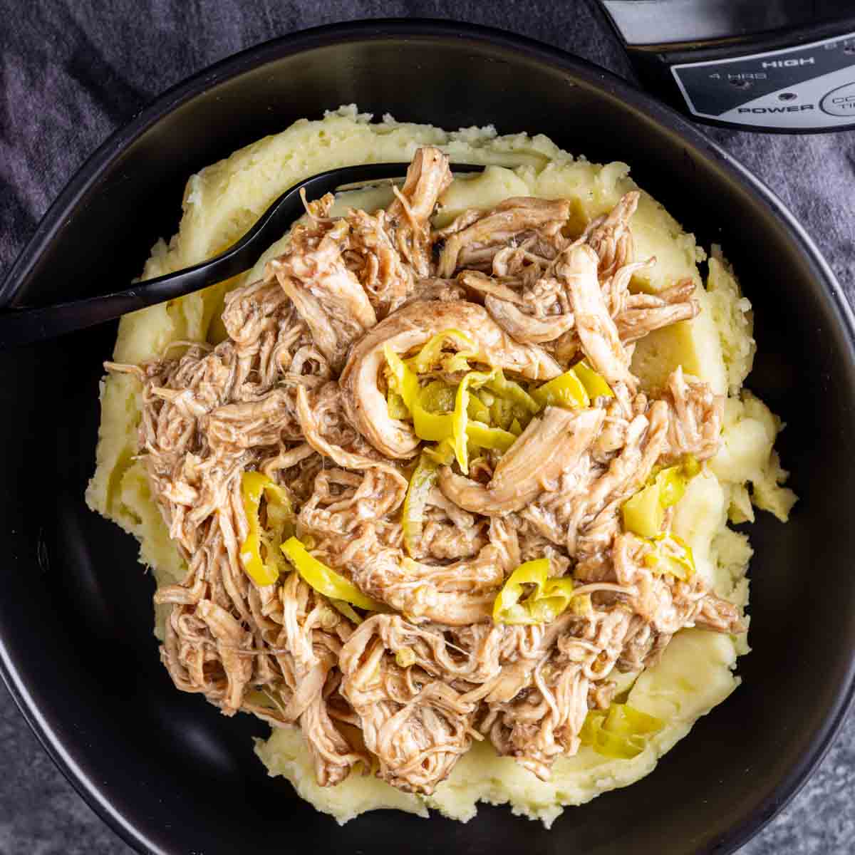 A black bowl filled with mashed potatoes topped with Slow Cooker Mississippi Chicken and sliced yellow peppers. A black spoon rests in the bowl.
