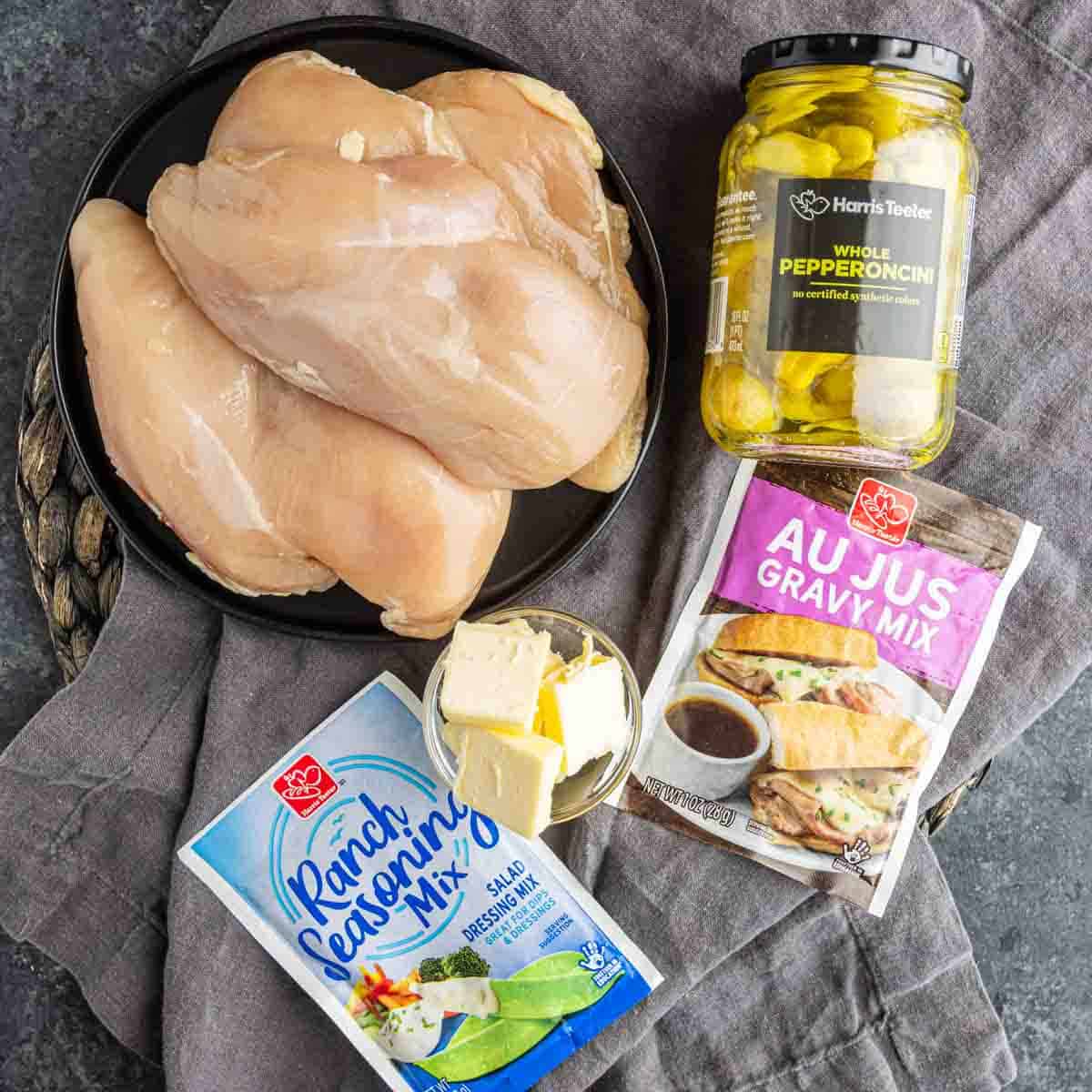 Raw chicken breasts, whole pepperoncini, ranch seasoning, au jus gravy mix packets, and butter on a plate—all you need for an easy dump and go crockpot meal like Slow Cooker Mississippi Chicken.