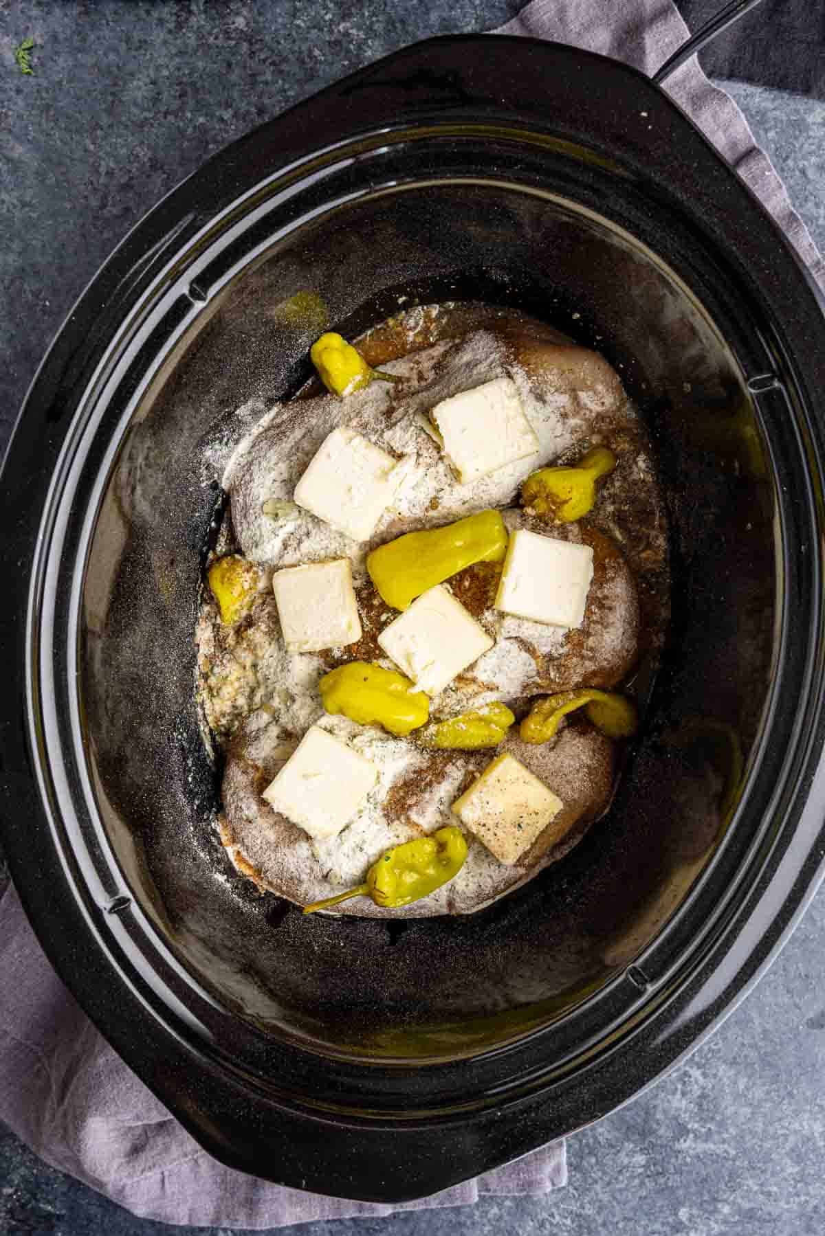 Three raw beef roasts in a slow cooker topped with sliced butter, pepperoncini peppers, and seasoning—an easy dump and go crockpot meal ready to be cooked.