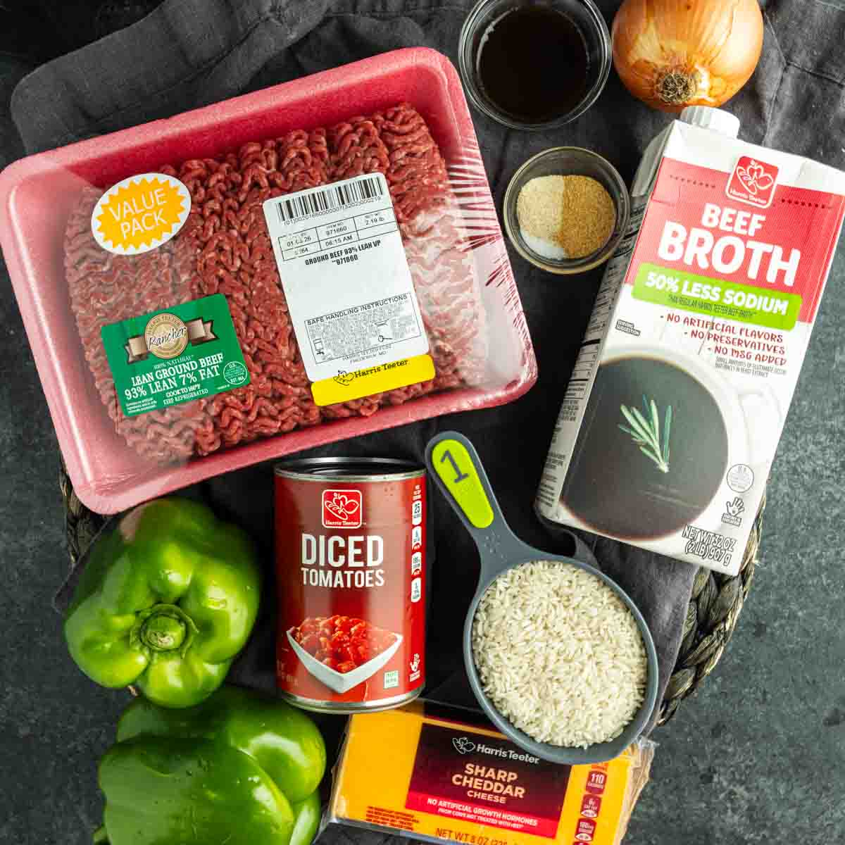 Top-down view of ingredients for a ground beef and rice skillet—beef broth, diced tomatoes, green bell peppers, cheddar cheese, onion, and seasonings—arranged on a dark surface for an easy unstuffed pepper skillet.