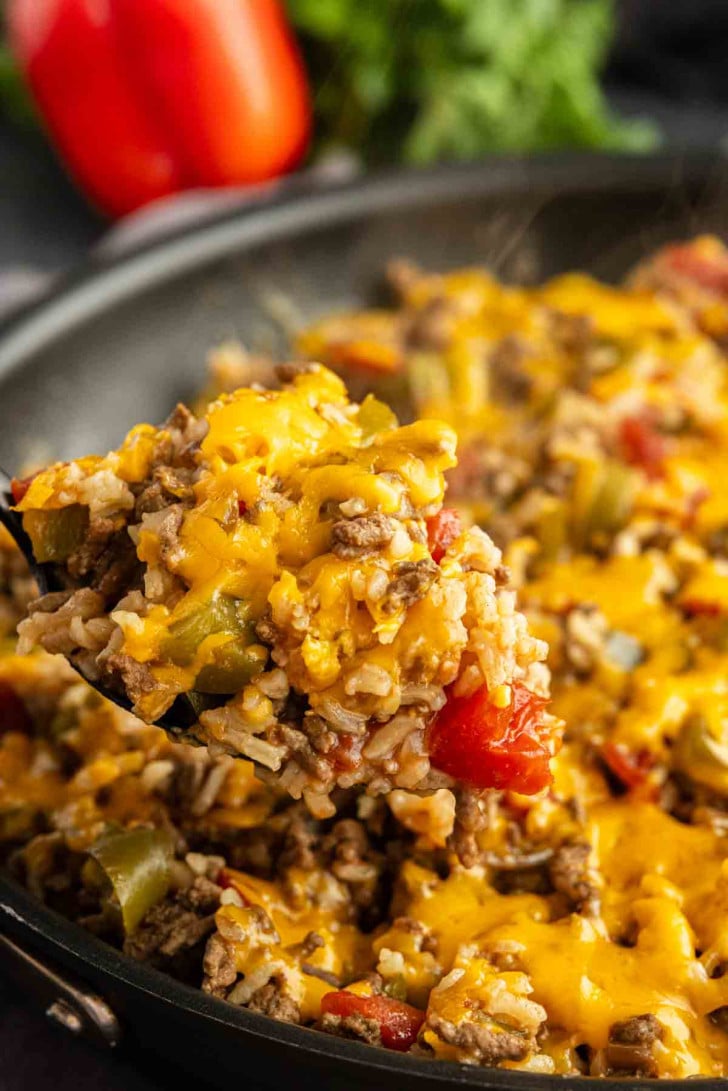 A close-up of a spoonful of cheesy ground beef, rice, diced tomatoes, and bell peppers being served from a skillet.