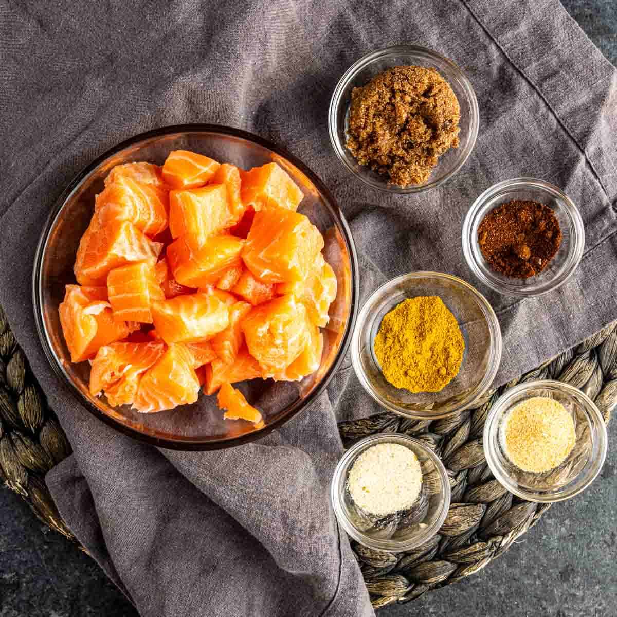 A bowl of raw salmon cubes, perfect for a crispy salmon bites recipe, sits beside small glass bowls of brown sugar, paprika, curry powder, onion powder, and garlic powder on a gray cloth.