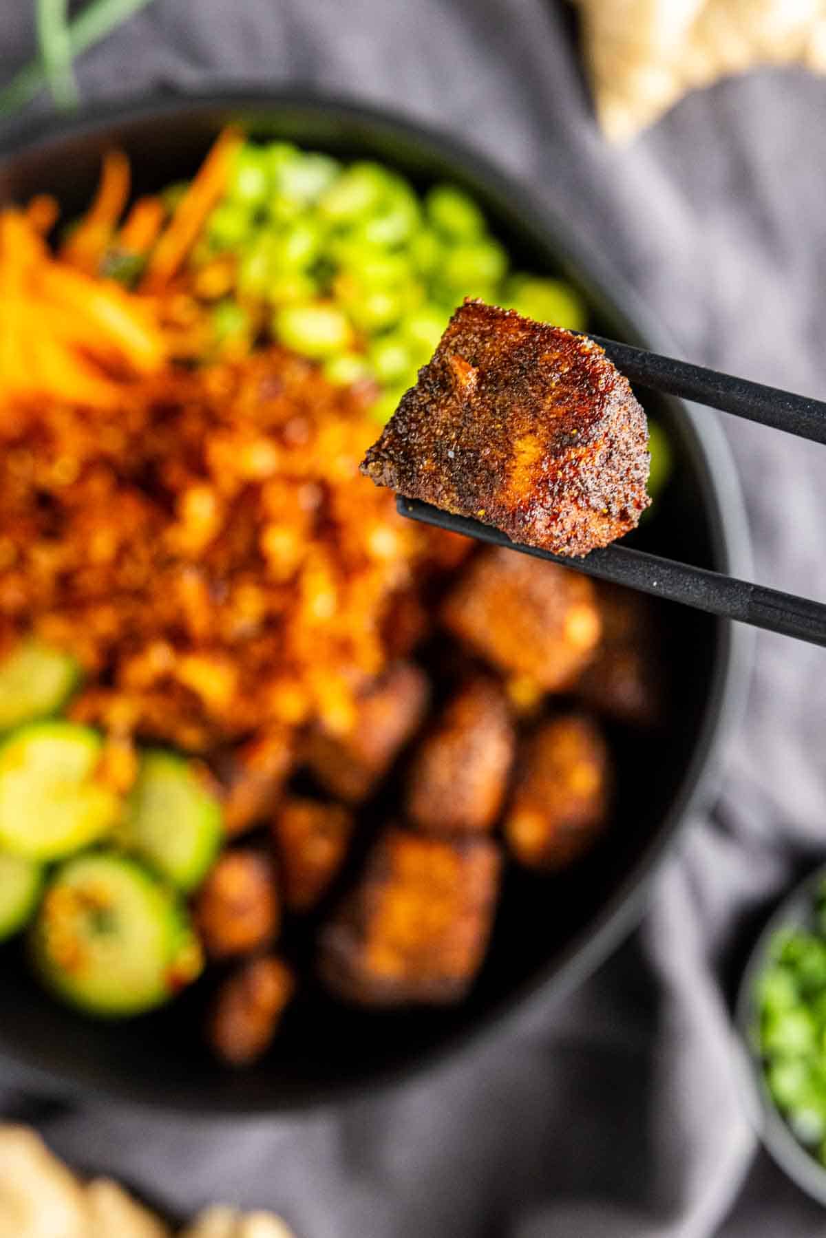 A close-up of chopsticks holding a piece of seasoned tofu above a bowl filled with tofu, sliced cucumbers, shredded carrots, edamame, and crispy toppings—perfect inspiration for your next salmon rice bowl or air fryer salmon bites recipe.