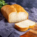 A loaf of white bread with a single slice cut, placed on a gray cloth on a wooden surface.