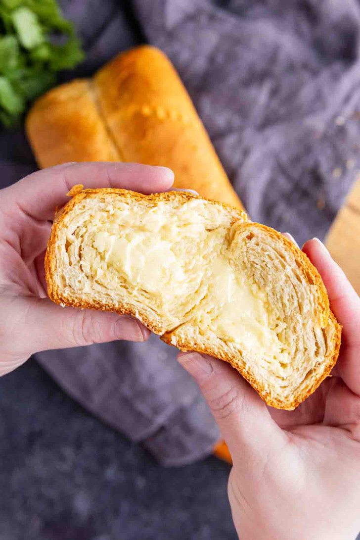 Two hands hold a crescent roll bread split in half, revealing its flaky layers and creamy filling, with a buttery bread loaf and a gray cloth in the background.