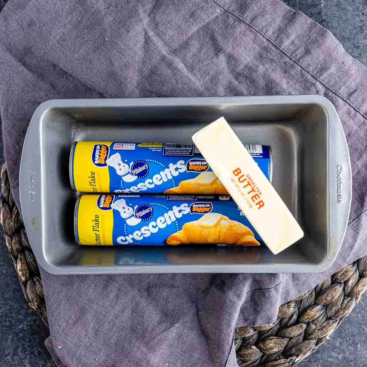 A metal baking pan with two cans of Pillsbury crescent rolls and a stick of butter inside, placed on a gray cloth.