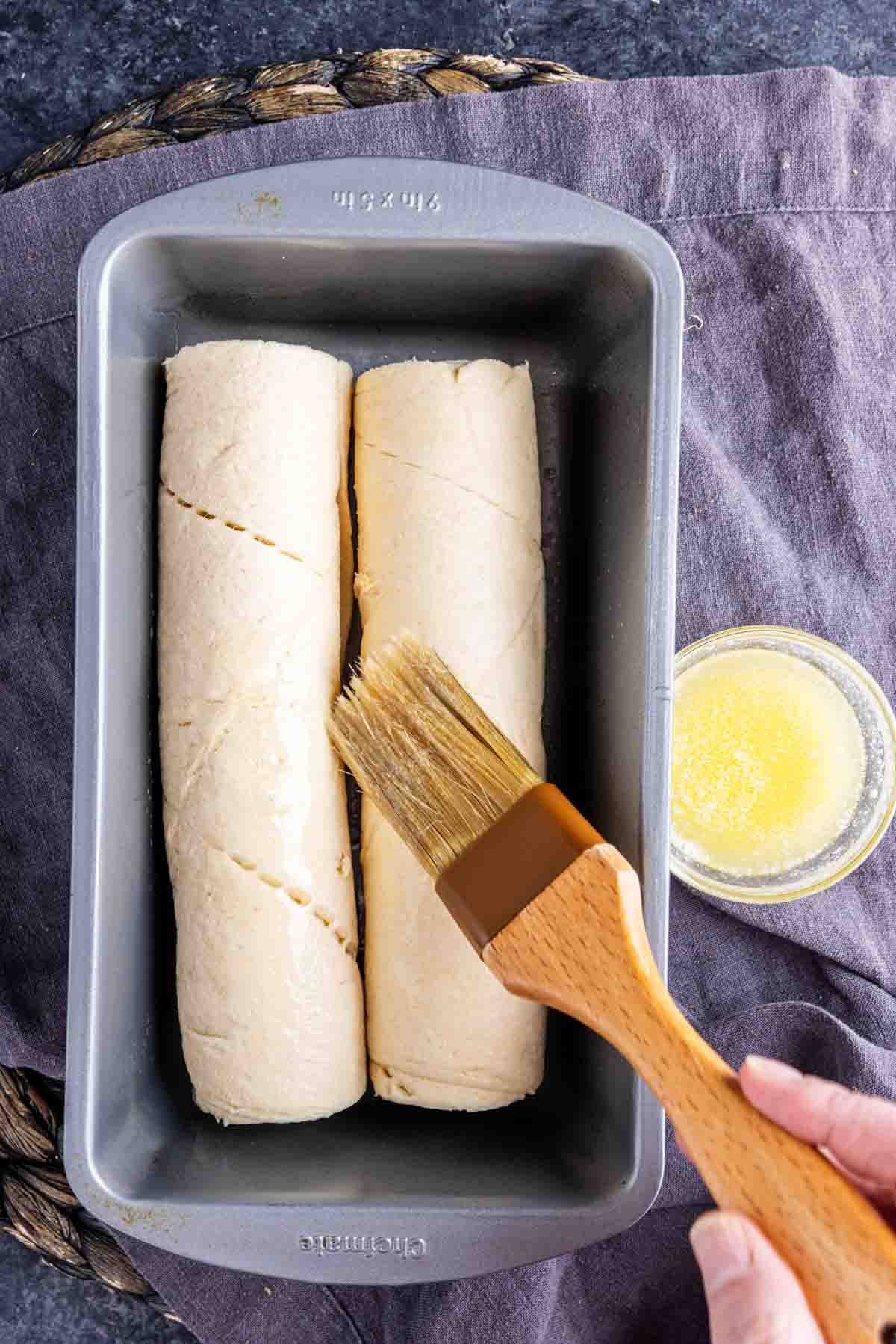 A hand uses a pastry brush to apply melted butter to two unbaked Viral TikTok Crescent Roll Loaf dough rolls in a loaf pan on a dark surface.