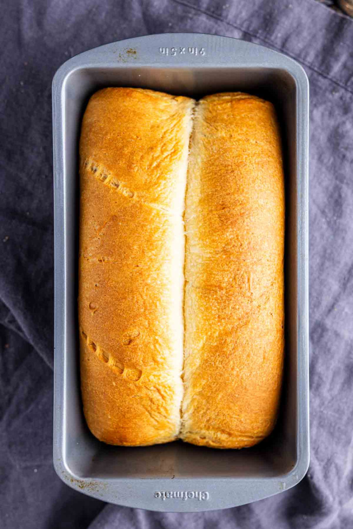 A loaf of golden brown bread in a rectangular metal baking pan, resting on a dark cloth.