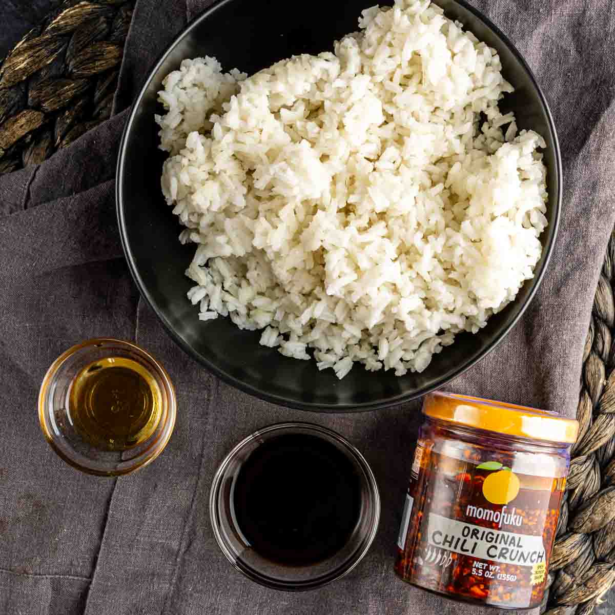 A bowl of cooked white rice next to a jar of Momofuku Chili Crunch, a small glass of oil, and a small bowl of soy sauce—perfect for making your own TikTok crispy rice or trying out a new crispy rice recipe.
