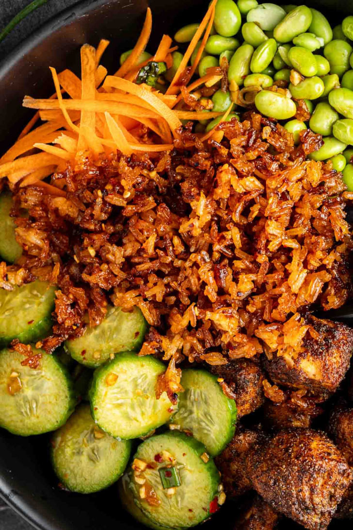A close-up of a delicious crispy rice bowl featuring shredded carrots, edamame, sliced cucumbers, and seasoned mushrooms.