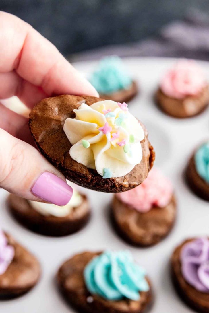 A hand with painted nails holds a small chocolate cookie topped with white frosting and pastel star sprinkles; more easy Easter dessert cookies made from brownie mix are on a plate in the background.