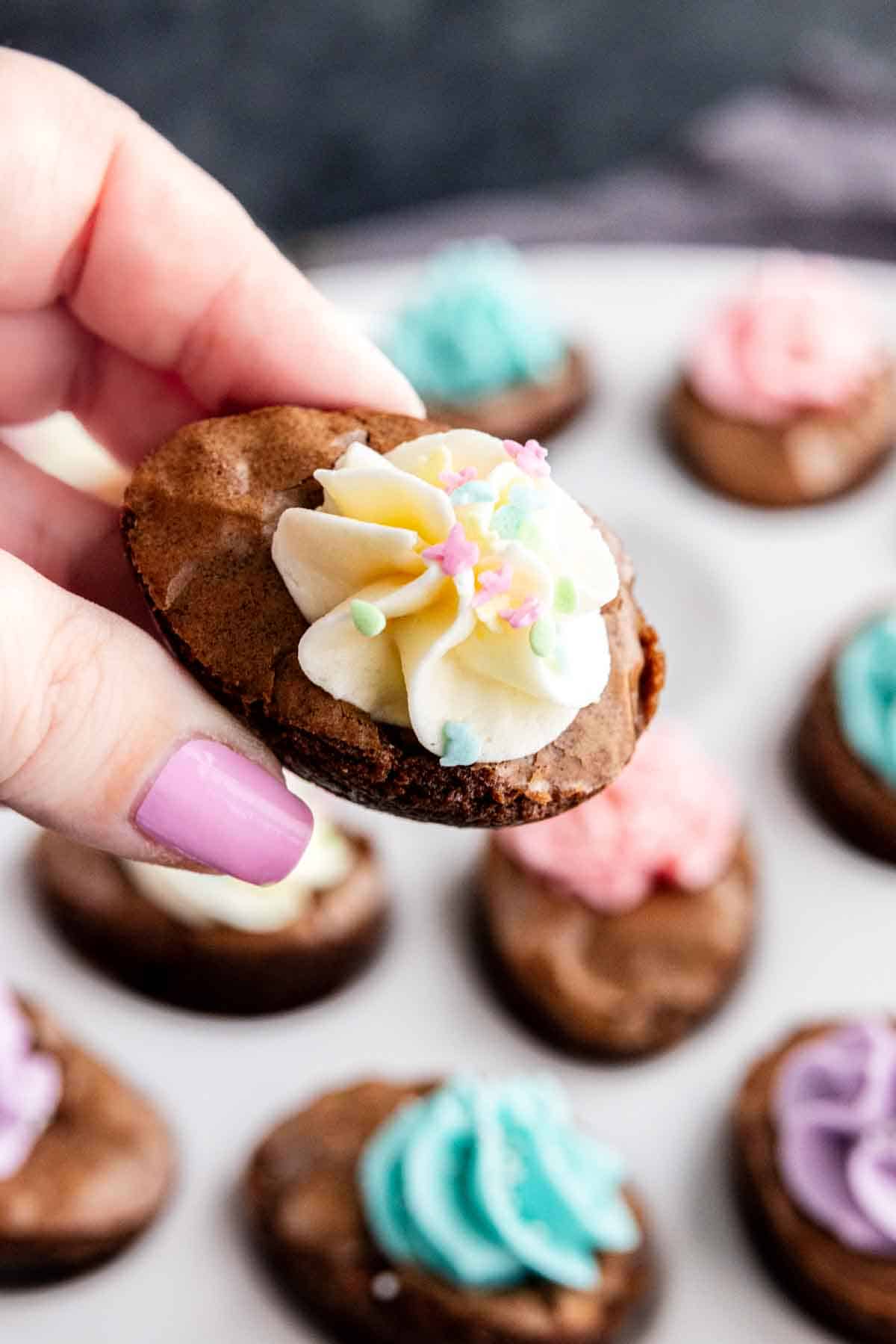 A hand with painted nails holds a small chocolate cookie topped with white frosting and pastel star sprinkles; more easy Easter dessert cookies made from brownie mix are on a plate in the background.