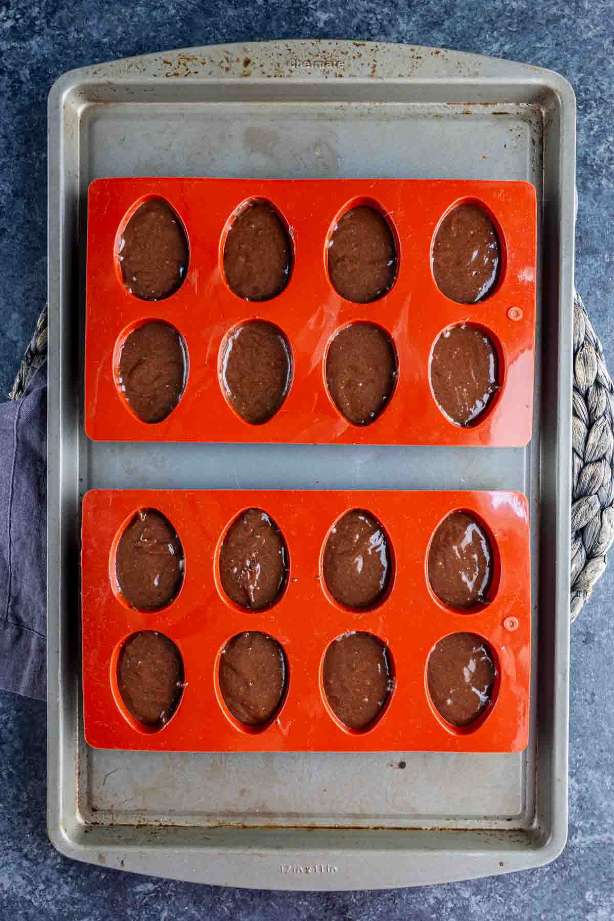 Two orange silicone egg molds filled with chocolate batter sit on a metal baking sheet, ready to become festive Easter brownies.