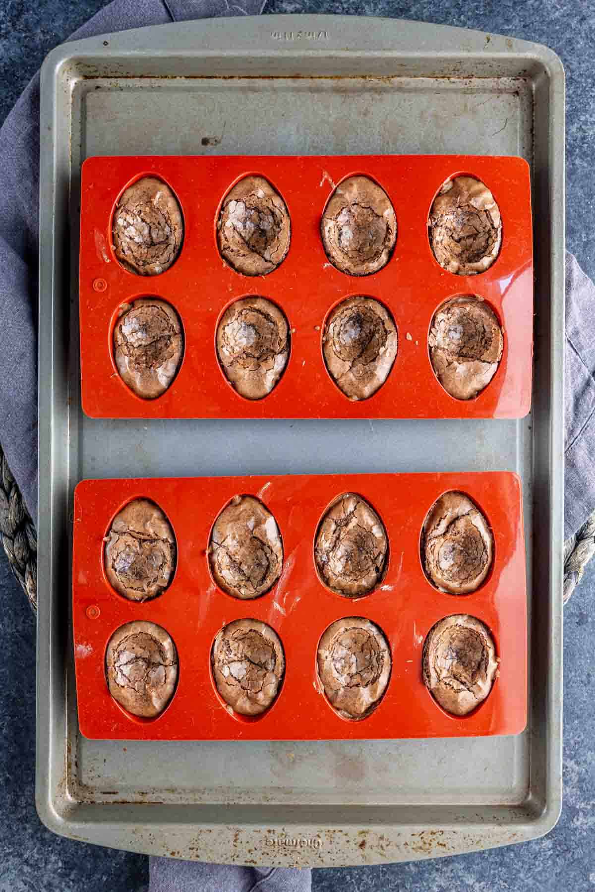 Two silicone baking molds filled with twelve chocolate, egg-shaped Deviled Brownies sit on a metal baking sheet—an easy Easter dessert perfect for spring parties.