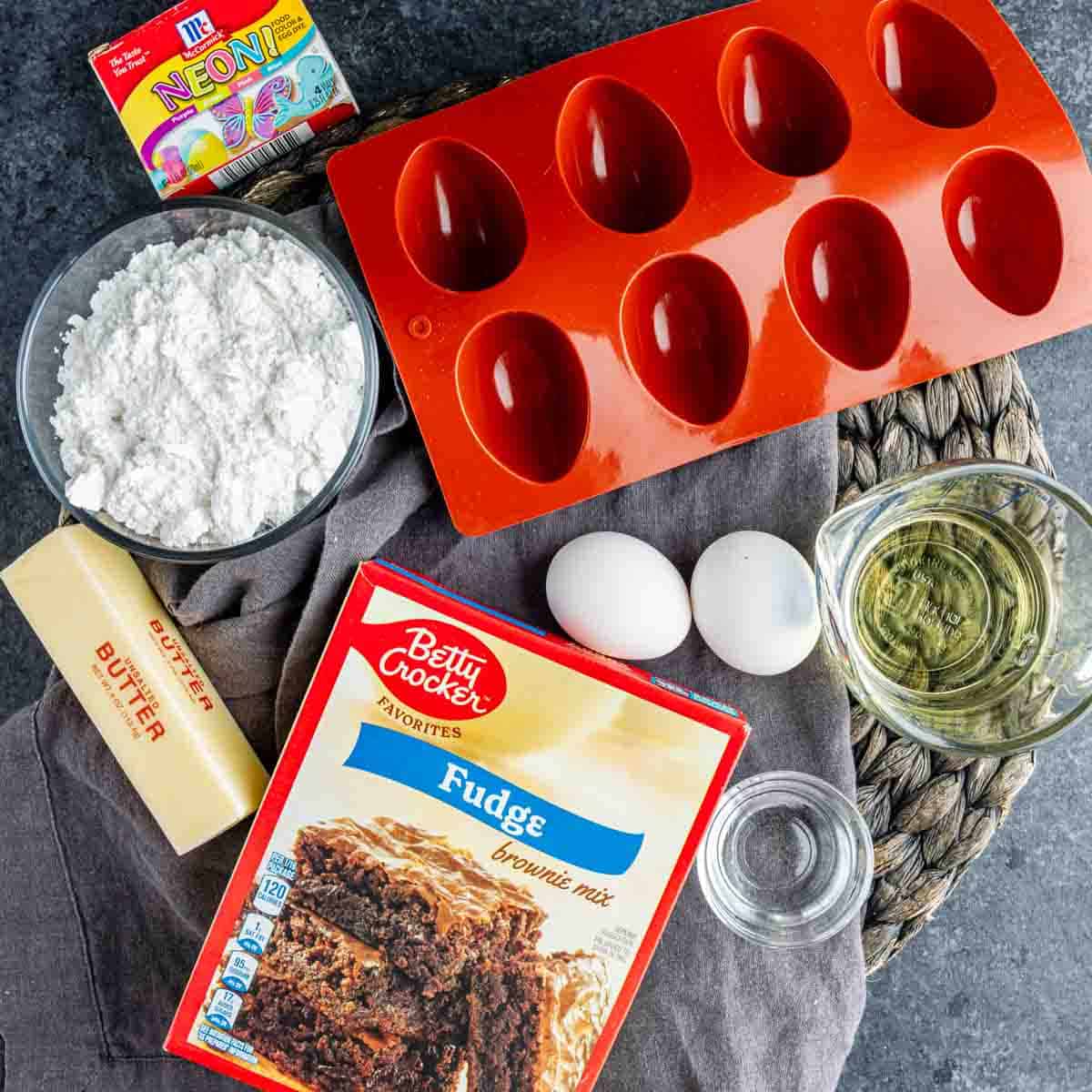 Brownie mix, butter, eggs, oil, water, powdered sugar, neon food coloring, and a red silicone egg-shaped mold arranged on a counter for easy Easter dessert Deviled Brownies.