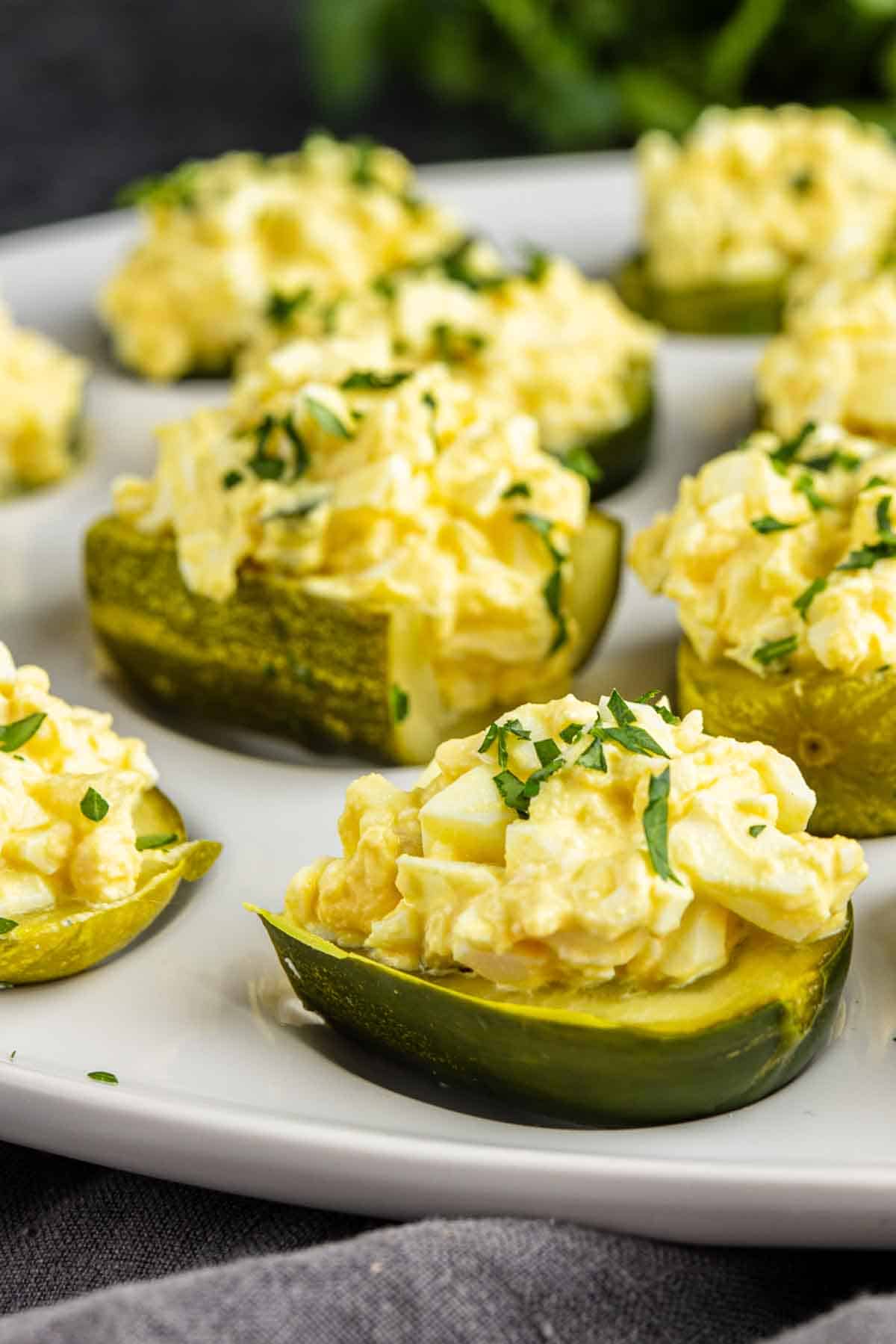 Close-up of sliced pickles topped with a creamy egg salad mixture and garnished with chopped herbs, these easy pickle appetizers are artfully arranged on a white plate.