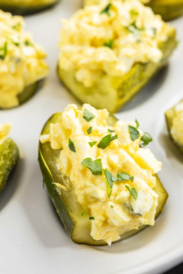 Halved pickles filled with chopped egg salad and garnished with parsley are arranged on a white plate.