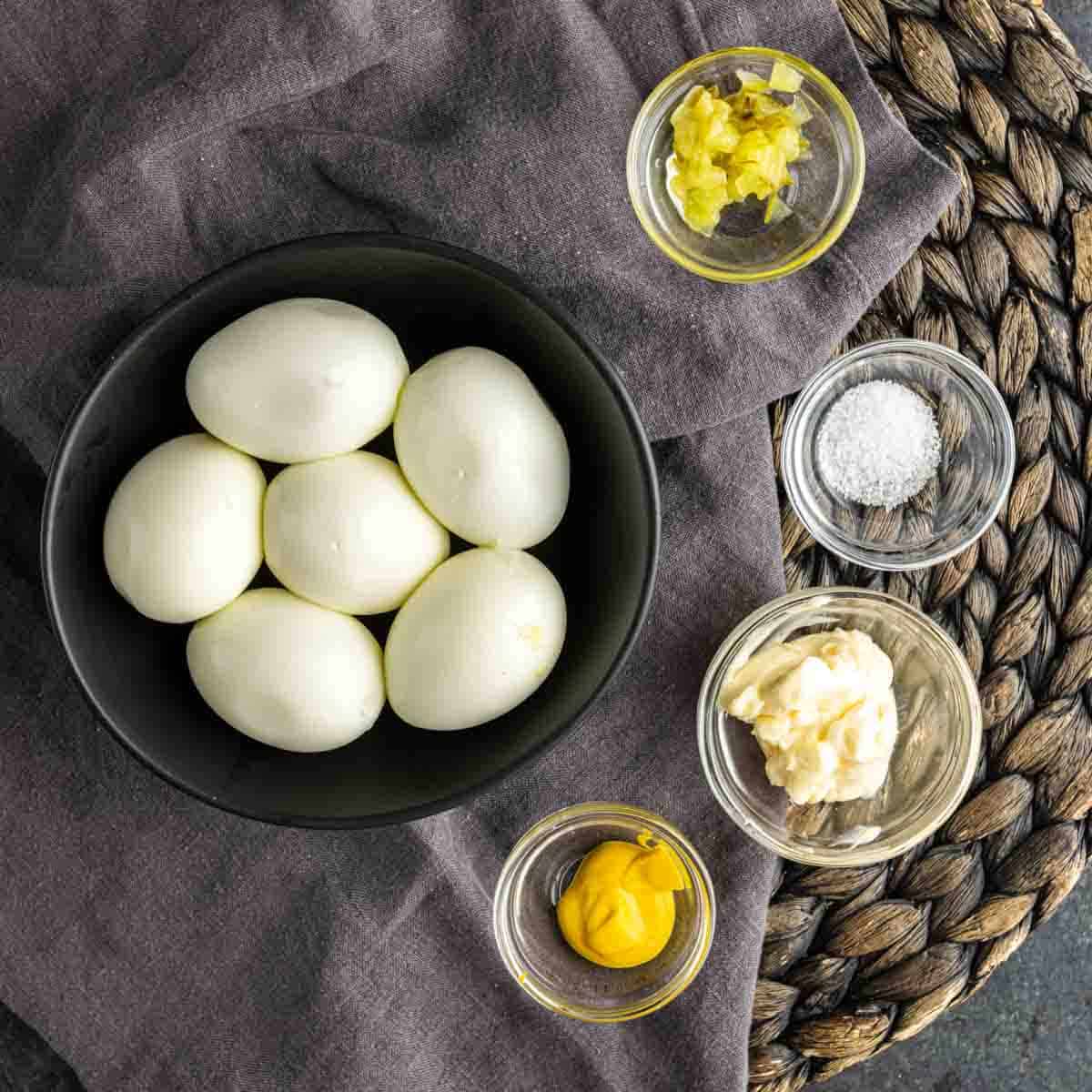 A black bowl with six hard-boiled eggs sits next to small glass bowls of chopped pickles, salt, mayonnaise, and yellow mustard—perfect for making deviled eggs with pickles or easy pickle appetizers on a gray cloth and woven placemat.